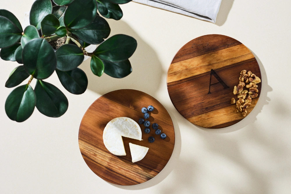 Bird's-eye view of two small round cutting boards for appetizers on a kitchen countertop, one featuring brie cheese, with a sunny ambiance and a plant nearby. One board is personalized with the letter 'A'.