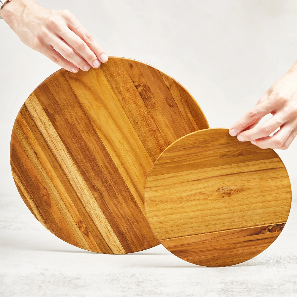Two round cutting boards, a small and a large one, held upright by two hands to showcase their main surfaces, made from 100% certified teak wood.