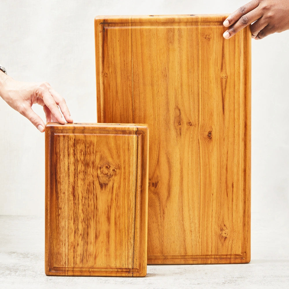 One large and one small premium teak cutting board with a juice groove and embedded handle, standing upright to display their main surfaces, made from 100% certified teak wood.