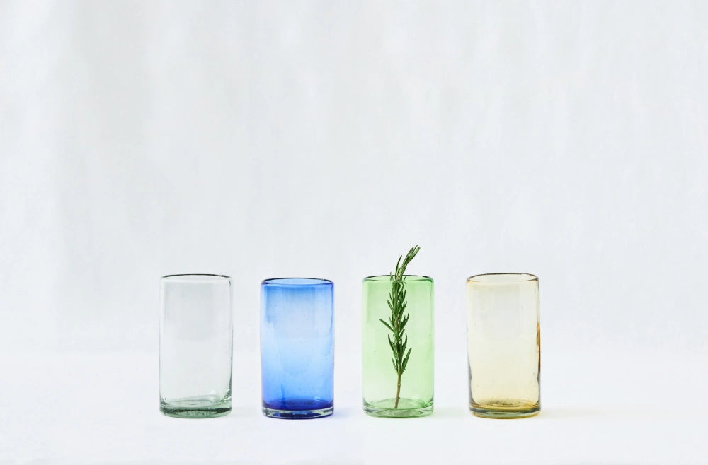 Four lightly tinted tall highball glasses in mixed colors, blue, green, amber and clear glass, made from 100% recycled glass, arranged in a row on a white background.
