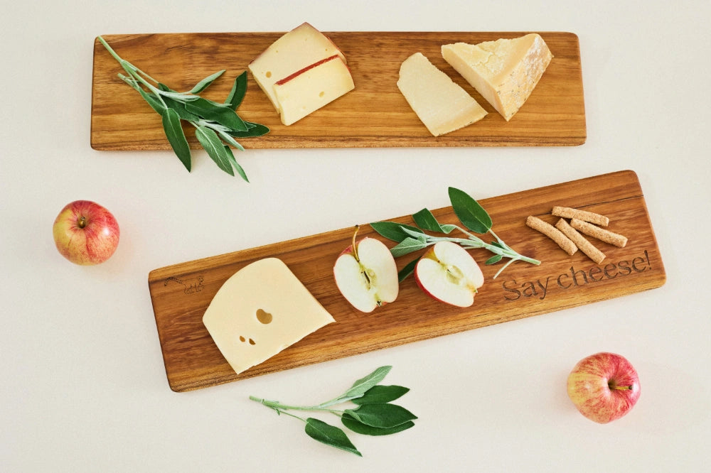 Two long, super lightweight teak cheeseboard trays with beveled edges, viewed from above on a kitchen counter, arranged with assorted cheeses, apple slices, and fresh sage leaves.