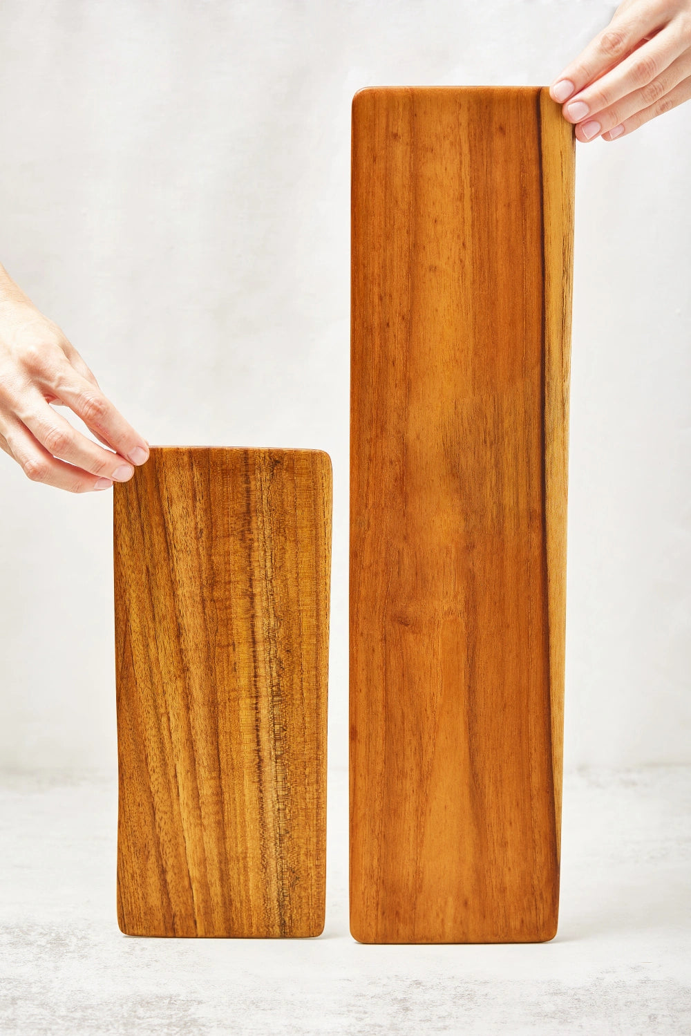 A short and a long teak cheeseboard tray, both standing upright, showcasing their smooth surfaces and beveled edges, made from 100% certified teak wood.