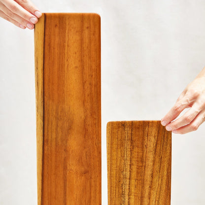 A short and a long teak cheeseboard tray, both standing upright, showcasing their smooth surfaces and beveled edges, made from 100% certified teak wood.