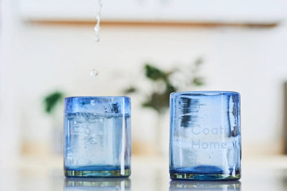 Two blue lightly tinted short glass tumblers, filled with water, arranged on a table in a housewarming setup, perfect for cocktails, made from 100% recycled glass.