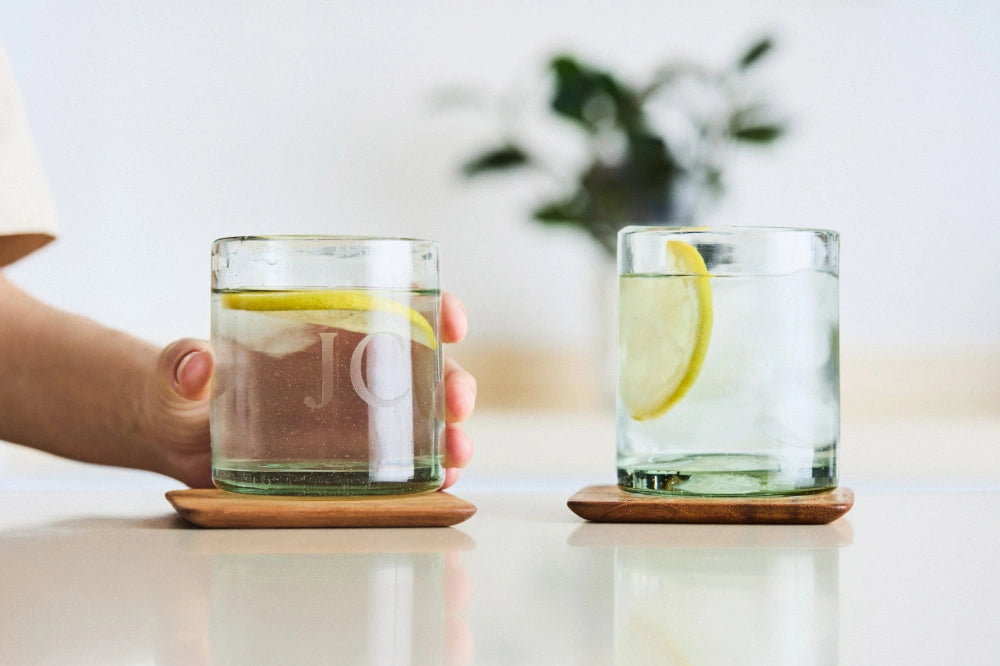 Two clear short glass tumblers, filled with water, arranged on a table in a housewarming setup, perfect for cocktails, made from 100% recycled glass.