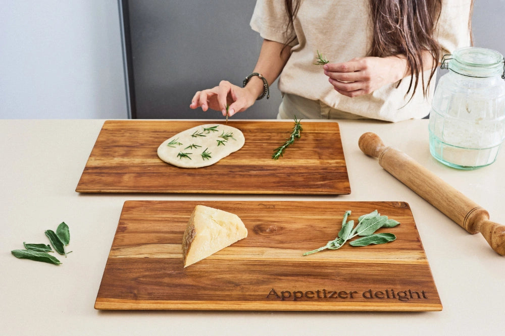 Two large, super lightweight teak cutting boards from a diagonal angle, one featuring a focaccia and the other showcasing Grana cheese, with one board personalized to say 'Appetizer Delight,' both with beveled edges on a kitchen counter.