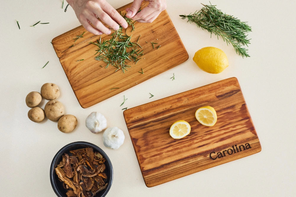 Two small, super lightweight teak cutting boards with beveled edges, personalized with a name, placed on a kitchen counter from a bird's-eye view, with rosemary, lemon, and other ingredients being prepared on them.