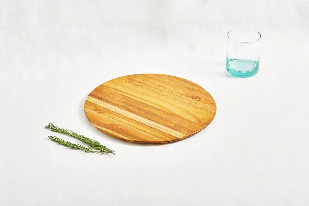 One large round cutting board made from 100% certified teak wood, with a short turquoise drinking glass and two sprigs of rosemary placed next to it, displayed on a white background.