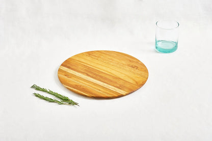 One large round cutting board made from 100% certified teak wood, with a short turquoise drinking glass and two sprigs of rosemary placed next to it, displayed on a white background.