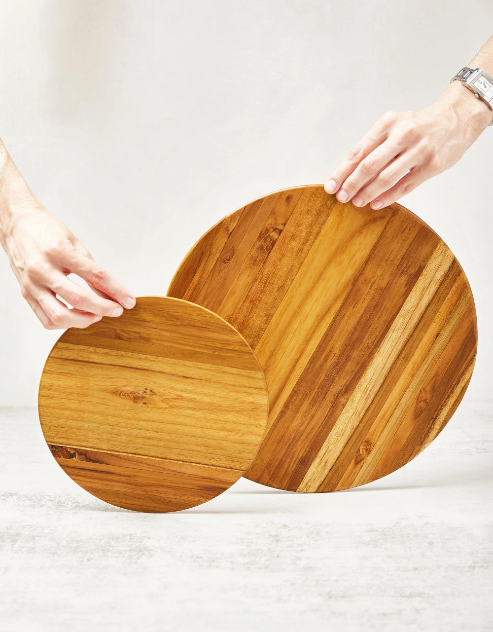 Two round cutting boards, a small and a large one, held upright by two hands to showcase their main surfaces, made from 100% certified teak wood.