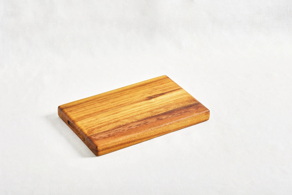 One small premium reversible teak cutting board, lying flat at a diagonal angle on a white background, highlighting its dual-use design and smooth surface, made from 100% certified teak wood.