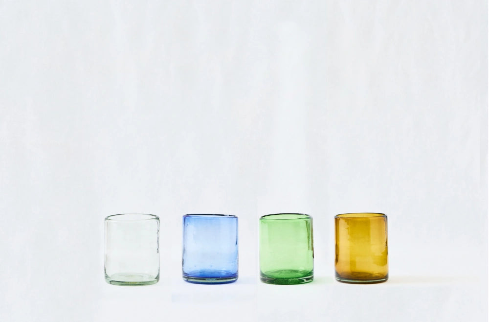 Four lightly tinted short glass tumblers, in clear, blue, green & amber glass, made from 100% recycled glass, arranged on a white background.