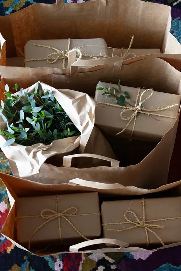 Three bags filled with eco-friendly gift items, accompanied by a bouquet of natural leaves, all showcasing sustainable packaging made from craft paper and tied with natural rope.