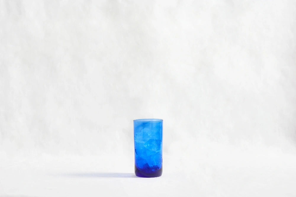 Blue deep tinted tall highball glass, made from 100% recycled glass, shown from the side on a white background.