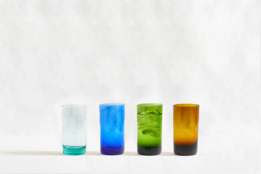 Four deep tinted tall highball glasses in mixed colors, made from 100% recycled glass, standing side by side on a white background.