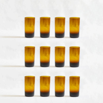 Twelve amber deep tinted tall highball glasses, made from 100% recycled glass, arranged together on a white background.