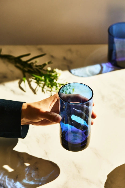 Blue deep tinted tall highball glass, filled with water, placed on a table in a summer party setup, made from 100% recycled glass.