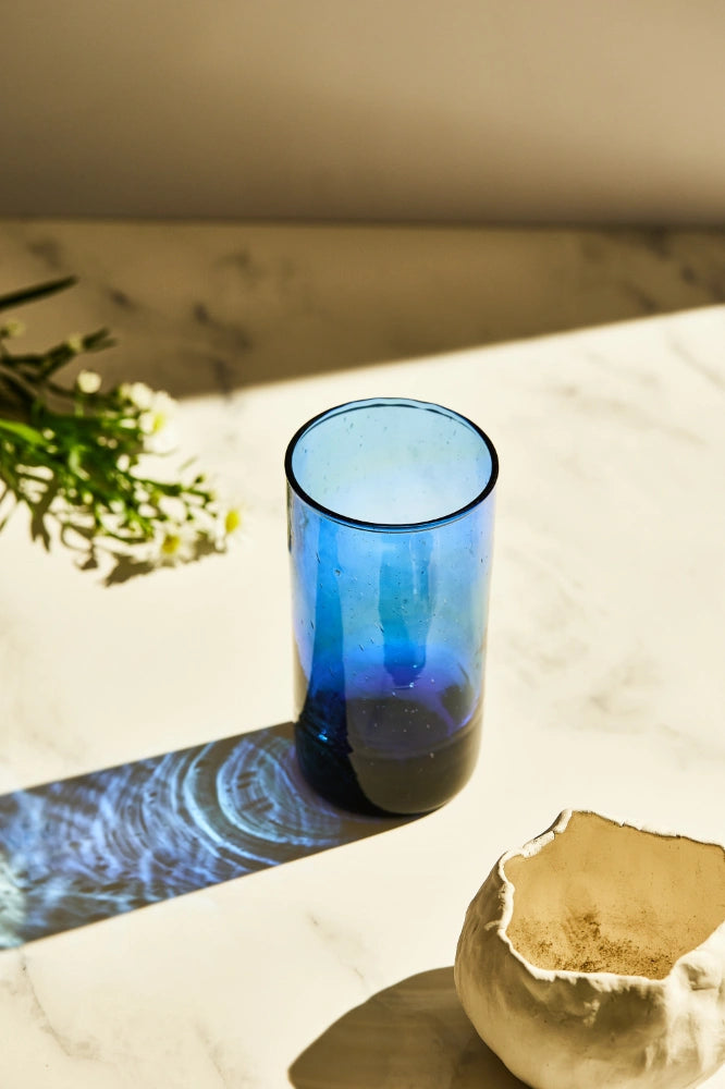 Blue deep tinted tall highball glass, filled with water, placed on a table in a summer party setup, made from 100% recycled glass.