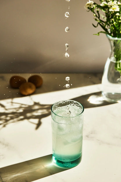 Turquoise tall highball glass, filled with water, placed on a table in a summer party setup, made from 100% recycled glass.