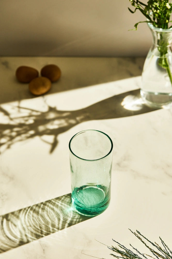 Turquoise tall highball glass, filled with water, placed on a table in a summer party setup, made from 100% recycled glass.